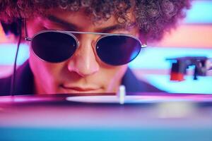 Young man with curly hair wearing sunglasses is focused on a vinyl record player in a vibrant atmosphere, showcasing the art of DJing and music mixing photo