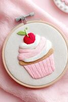 Embroidered cupcake design featuring pink frosting, a cherry on top, and a textured background, showcasing intricate craftsmanship and artistic flair in a circular hoop photo