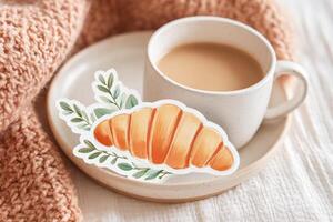Cozy setting featuring a cup of coffee beside a decorative croissant sticker on a plate, surrounded by a soft knitted blanket, creating a warm and inviting atmosphere for relaxation photo