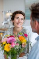 Smiling woman with a bouquet of colorful flowers, sitting in a bright room, engaging in conversation, radiating warmth and joy in a cozy atmosphere of connection and happiness photo