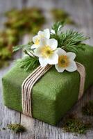 Green gift box wrapped in natural twine, adorned with white flowers and greenery, resting on a rustic wooden surface surrounded by moss, conveying a sense of nature and celebration photo