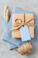Gift box wrapped in brown paper with twine, accompanied by a blue tag, placed on soft blue fabric, with decorative wheat stalks and twine, creating a cozy, rustic atmosphere photo