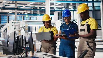 All black inspectors team in photovoltaics factory look over documents, review performance of robot arms placing solar panels on assembly lines. Supervisors inspecting solar cells plant. Camera B. video
