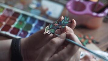 Person's hand coloring pin with branch with berries and flowers black aquarelle paint. Process of decorating brooch made of plywood with pattern applied by laser machine. Handmade and diy in workshop. video