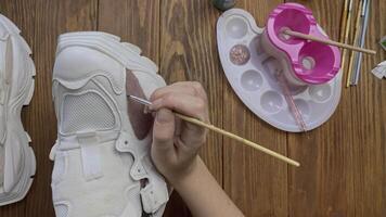 Flat lay of hands paint white boots with brush on wooden table. Hobby, creativity and handmade work at home. Designer's workshop. Palette, brown acrylic, bottle with water and other tools on desktop. video