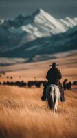 Cowboy rides a horse across golden grasslands with snow-capped mountains in the background during sunset photo
