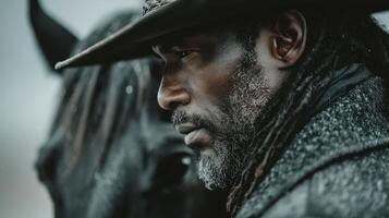 Man with beard and long hair wearing hat stands beside dark horse in an atmospheric outdoor setting photo