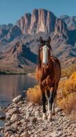 Majestic horse stands along a riverbank with towering mountains in the background under a clear blue sky photo