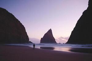 A person standing on the beach near a large rock photo