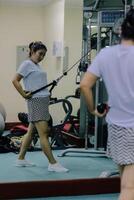 A woman indonesia working out at a gym. She uses a weight machine with a rope, pulling on the handle. Her reflection is seen in a large mirror in front of her. photo