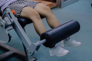 close up of a woman's legs using a leg extension machine, sitting on a cushioned chair with her legs propped up behind a dark cushioned bar that is part of the machine photo