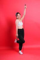A woman in fitness attire is holding a jump rope, standing on one leg, isolated on a red background. photo