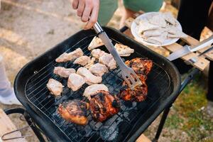 Chef turning over pieces of marinated chicken and pork on small barbecue grill during summer garden party, with plate of flatbread wraps in background. Person grilling marinated chicken and pork photo