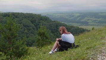 Young woman sits on a grassy hillside, absorbed in the breathtaking view of lush green mountains. The serene landscape stretches into the distance under a cloudy sky video