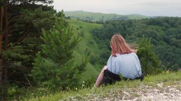 Young woman hiker sitting on the edge of a cliff enjoying the view of a green valley covered in trees, embracing the peace and quiet of nature during a summer trekking adventure video