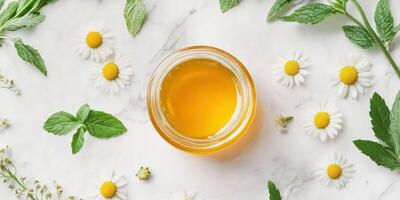 Flat lay of golden honey jar with fresh chamomile and mint leaves on marble surface photo