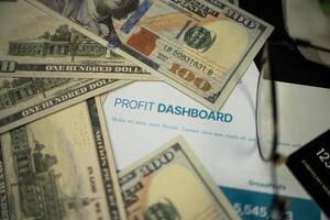 an image that focuses on a paper that says profit dashboard surrounded by several dollar bills, credit cards, and a pair of sunglasses on a table. Business and financial purposes photo