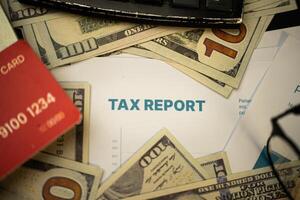 an image that focuses on a paper that says TAX REPORT surrounded by several dollar bills, credit cards, a calculator and a pair of sunglasses on a table. Business and financial purposes photo