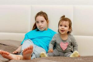 two sisters, older and younger, emotionally watch TV while sitting on the bed with the remote control in their hands photo