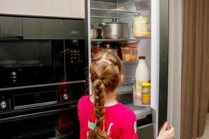a little girl stands at the open refrigerator door and thinks what products to take photo