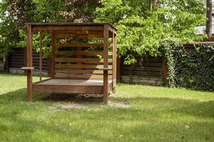 Wooden brown gazebo with a canopy in the middle of the garden. Picnic gazebo among green trees, surrounded by a fence with ivy, on a bright lawn. photo