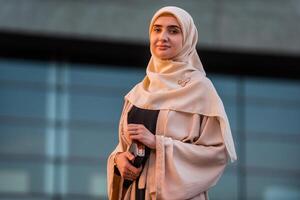 Portrait of a young confident Muslim business woman in hijab near the modern office building, looking at the camera with a friendly smile. photo