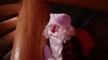 Santa Claus hiding in a wooden house. Authentic Santa with white beard and red costume with a sack with presents looking out of wooden pillar. video