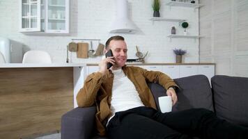 Cheerful young man talking the phone on sofa. Guy is laying on couch with a cup of coffee and having a pleasant conversation through mobile phone at home. video