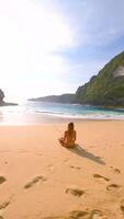 FPV drone shot of woman meditating alone on remote tropical beach with turquoise sea, waves, and cliffs, Bali, Indonesia. Peaceful scene of relaxation, mindfulness, and inner peace video