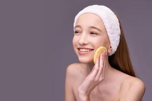 Young caucasian female using facial sponge for skincare routine with a smile on gray background. photo
