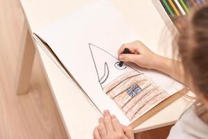 Young caucasian child drawing house with crayons on paper at table in bright room. photo