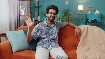 Indian man smiling friendly at camera, waving hands gesturing hello, hi, greeting at home on sofa video