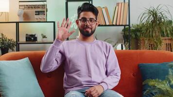 Indian man smiling friendly at camera, waving hands gesturing hello, hi, greeting at home on sofa video