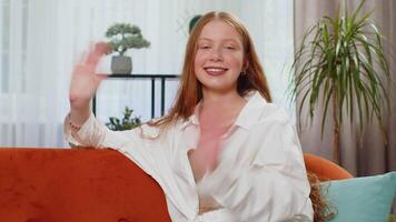 Pretty girl smiling friendly at camera, waving hands gesturing hello, hi, greeting at home on sofa video