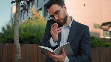 Busy Caucasian adult businessman talking with client mobile phone conversation writing notes multitasking man male manager working outdoors write schedule in notebook book business consulting in city video