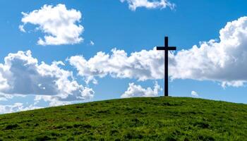 Silhouette of a cross atop a green hill beneath a bright blue sky and soft white clouds. A peaceful, spiritual scene ideal for Ascension Day, faith, and Christian reflection. photo