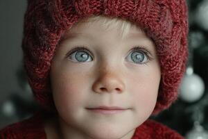 Child with bright blue eyes wearing red knitted hat looking thoughtfully at a decorated Christmas tree in winter photo