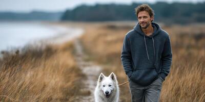 Man walking with a white dog along a grassy path near a serene lake during a cloudy afternoon photo