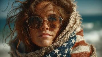 Woman wrapped in an American flag blanket at the beach on a sunny day with windswept hair and sunglasses photo