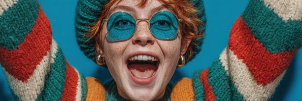 Young woman with playful smile in colorful sweater and retro sunglasses celebrates in front of a vibrant blue backdrop photo