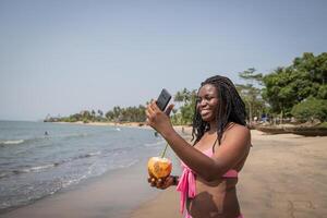 Charming african lady taking a selfie on the beach while enjoying coconut milk photo