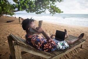 The digital vacationer lying on the bench by the sea makes a call while using his computer photo