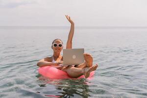 A woman is sitting on a pink inflatable raft in the ocean with a laptop on her lap. She is smiling and she is enjoying her time. photo