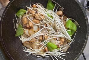 A frying pan filled with enoki mushrooms, chicken pieces, bell peppers, and onions. A colorful and appetizing dish in the process of cooking photo