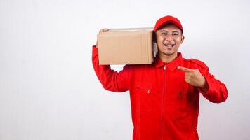 Delivery man in red uniform carrying stack of package box parcel on his shoulder and pointing at the package box isolated on white background photo