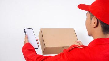 Rear view of delivery man in red uniform holding a package box parcel and smartphone isolated on white background photo