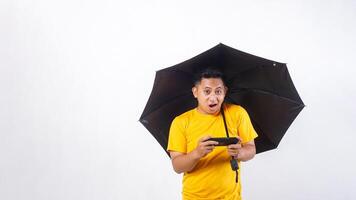 Asian man holds an umbrella while playing smartphone with shocked expression isolated on white background photo