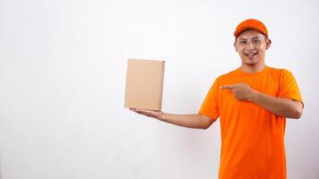 Asian deliveryman in orange t-shirt uniform with a package box parcel and pointing at it isolated on white background photo