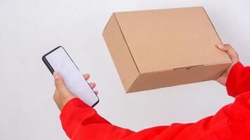 Close up of delivery man in red uniform holding a package box parcel and smartphone isolated on white background photo