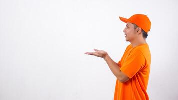 Side view of Asian deliveryman in orange t-shirt uniform poses as if he is presenting something in empty space area isolated on white background photo